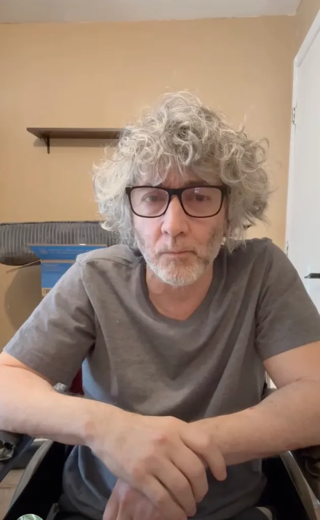 A man with curly gray hair and glasses looks directly at the camera, his hands clasped in front of him.